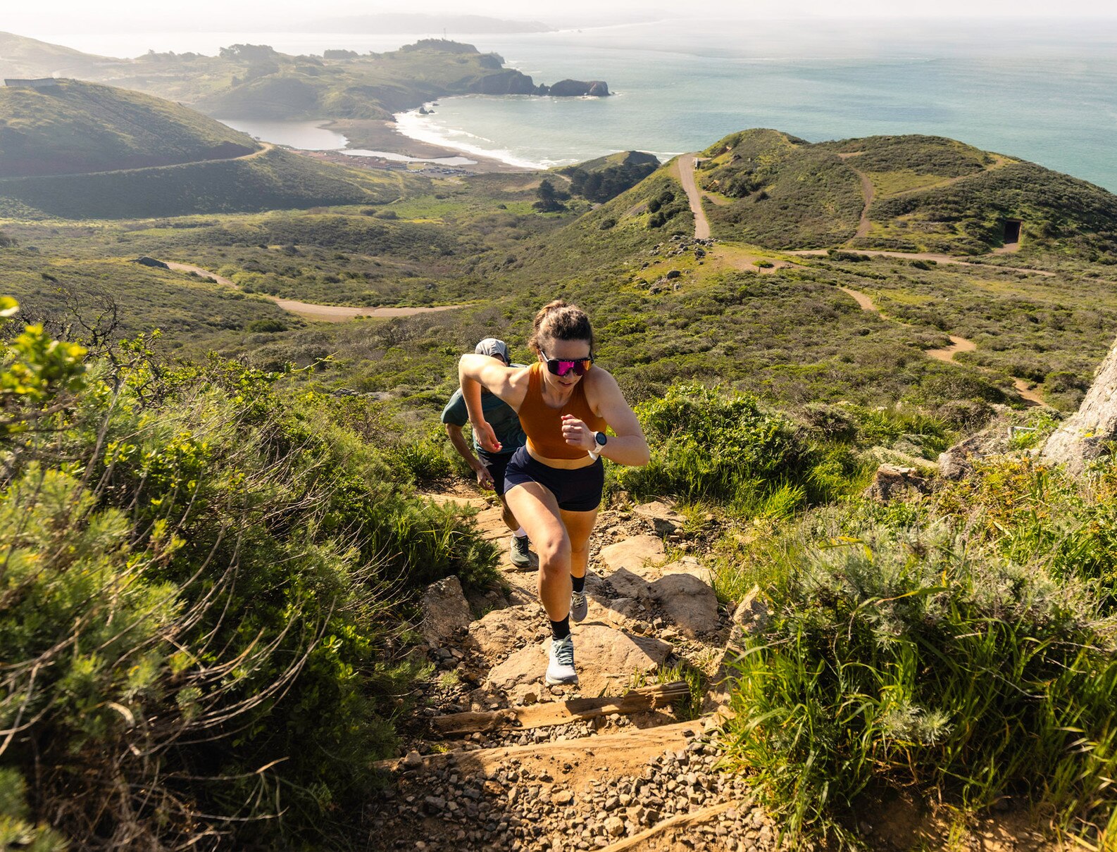 A female runner in black athletic wear and sunglasses runs uphill on a rugged coastal trail, with a scenic view of hills and ocean in the background