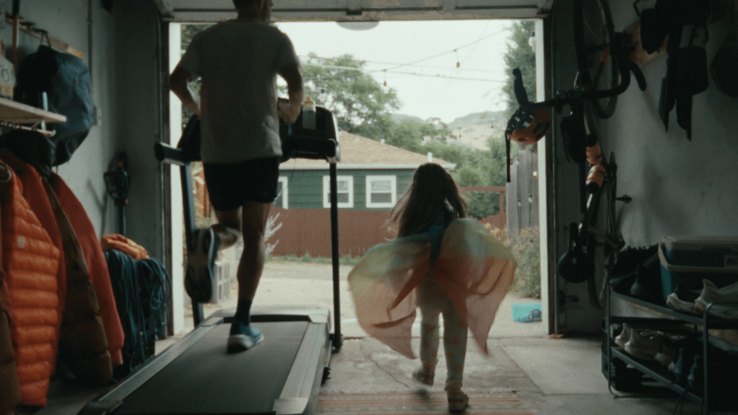 A runner trains on a treadmill in his garage as his child plays beside him.