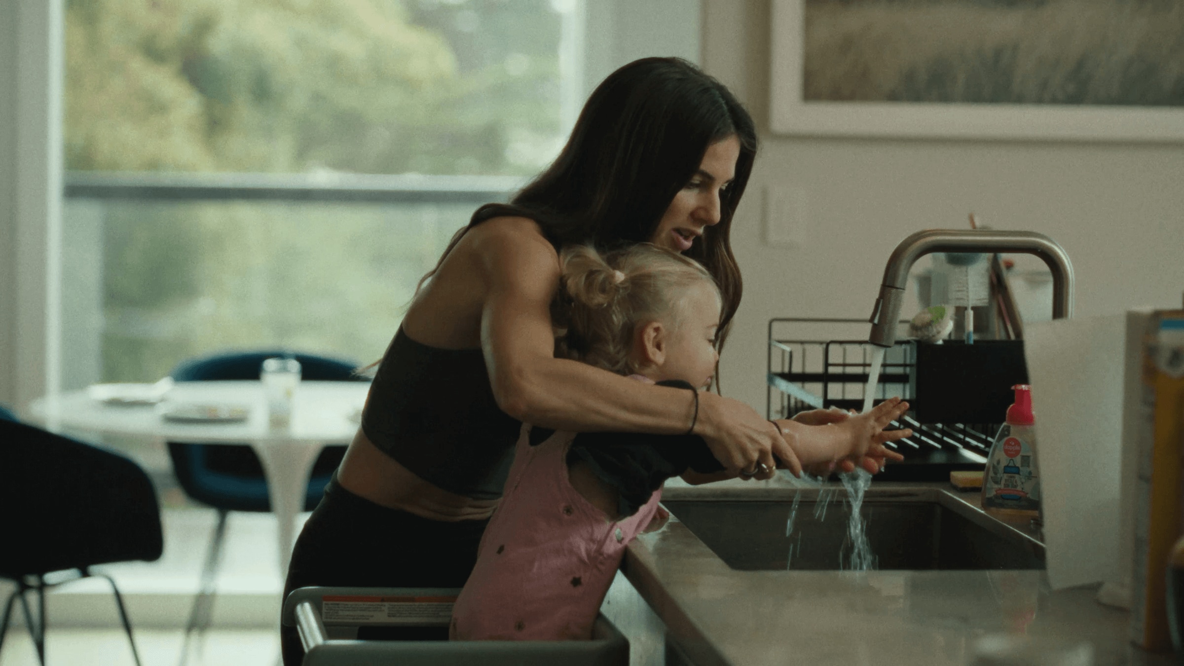 An adult helps a young child wash their hands at a kitchen sink, guiding the child's arms under the running water.