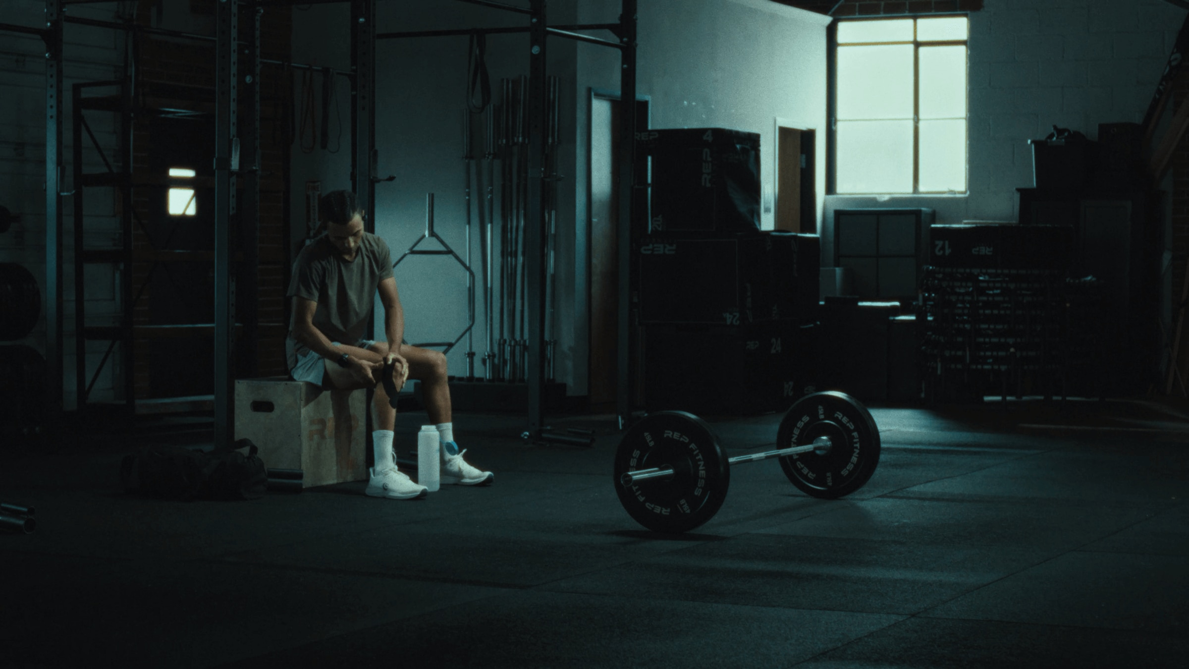 An athlete sits on a wooden box in a dimly lit gym, resting with a barbell on the floor in front of them.