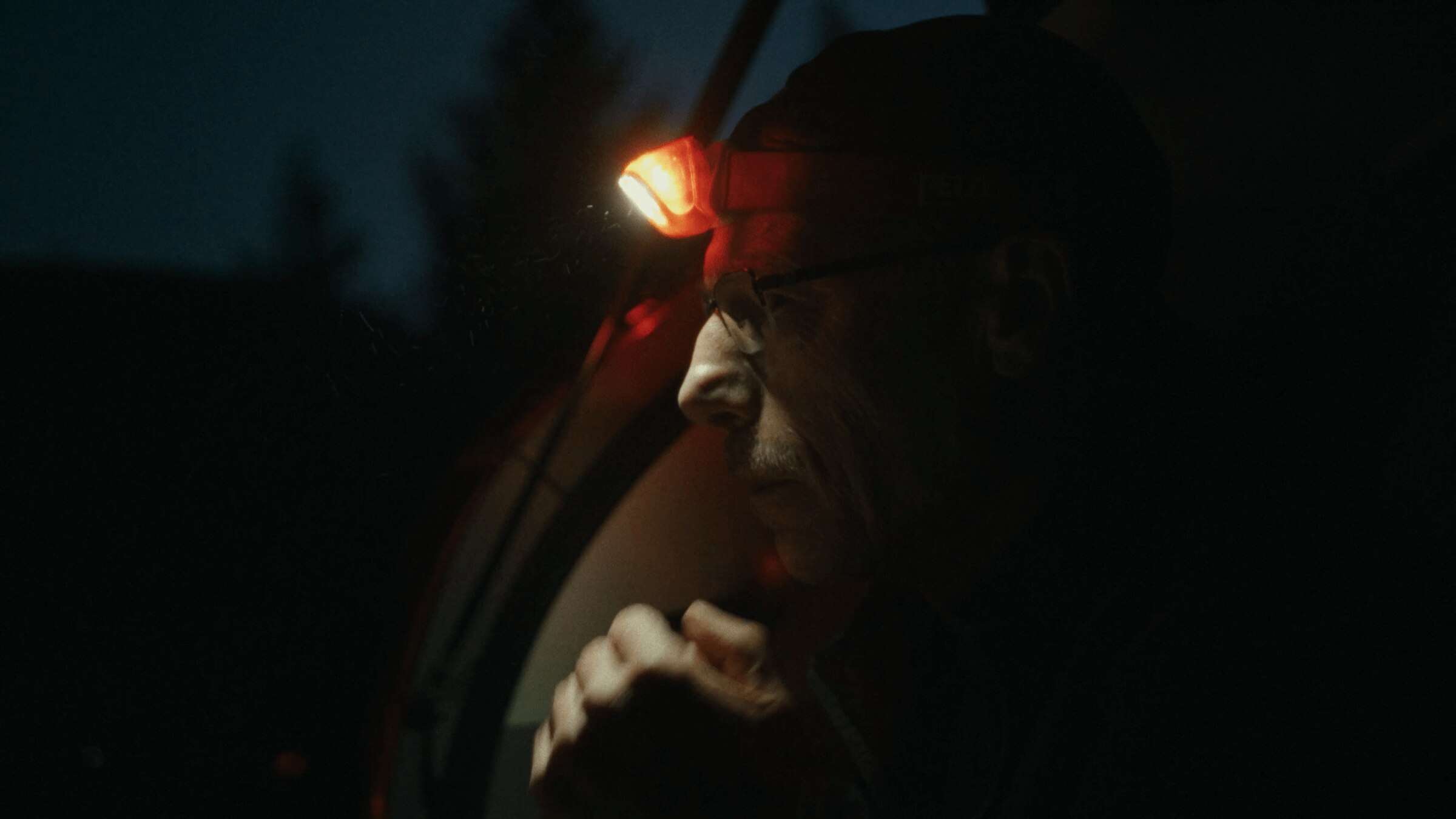 A person wearing a red headlamp sits in the dark outdoors, their face partially illuminated by the light as they look ahead thoughtfully.